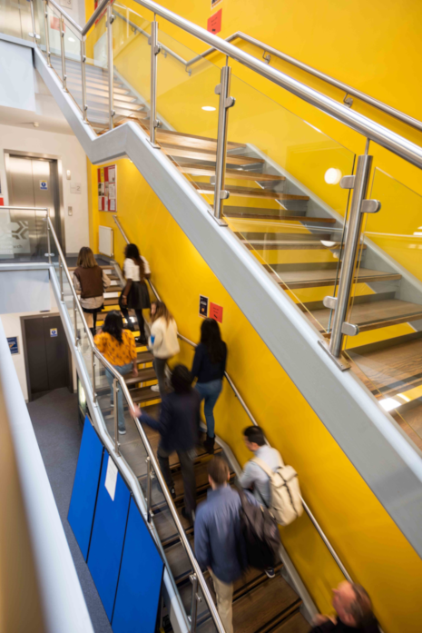 An indoor setting, an educational building. A bright yellow wall serves as the backdrop for a staircase with silver handrails and glass balustrades. Multiple individuals are seen ascending or descending the stairs, each with distinct attire. The floor appears to be carpeted, and the overall ambiance suggests a bustling environment.