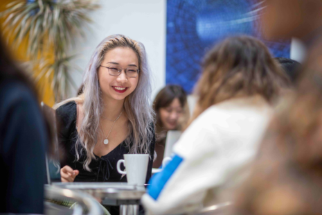 An indoor setting, possibly a cafe or a casual meeting place. A young woman with long, silver-coloured hair and glasses is prominently featured in the foreground. She is wearing a black top and has a necklace with a pendant. She is smiling and seems to be engaged in a conversation with someone out of the frame. In the background, there are other individuals, but their features are not distinctly visible. A white mug is placed on a table in front of the woman, suggesting she might be having a beverage. The ambiance is relaxed, with a hint of modernity, evident from the contemporary artwork on the wall.