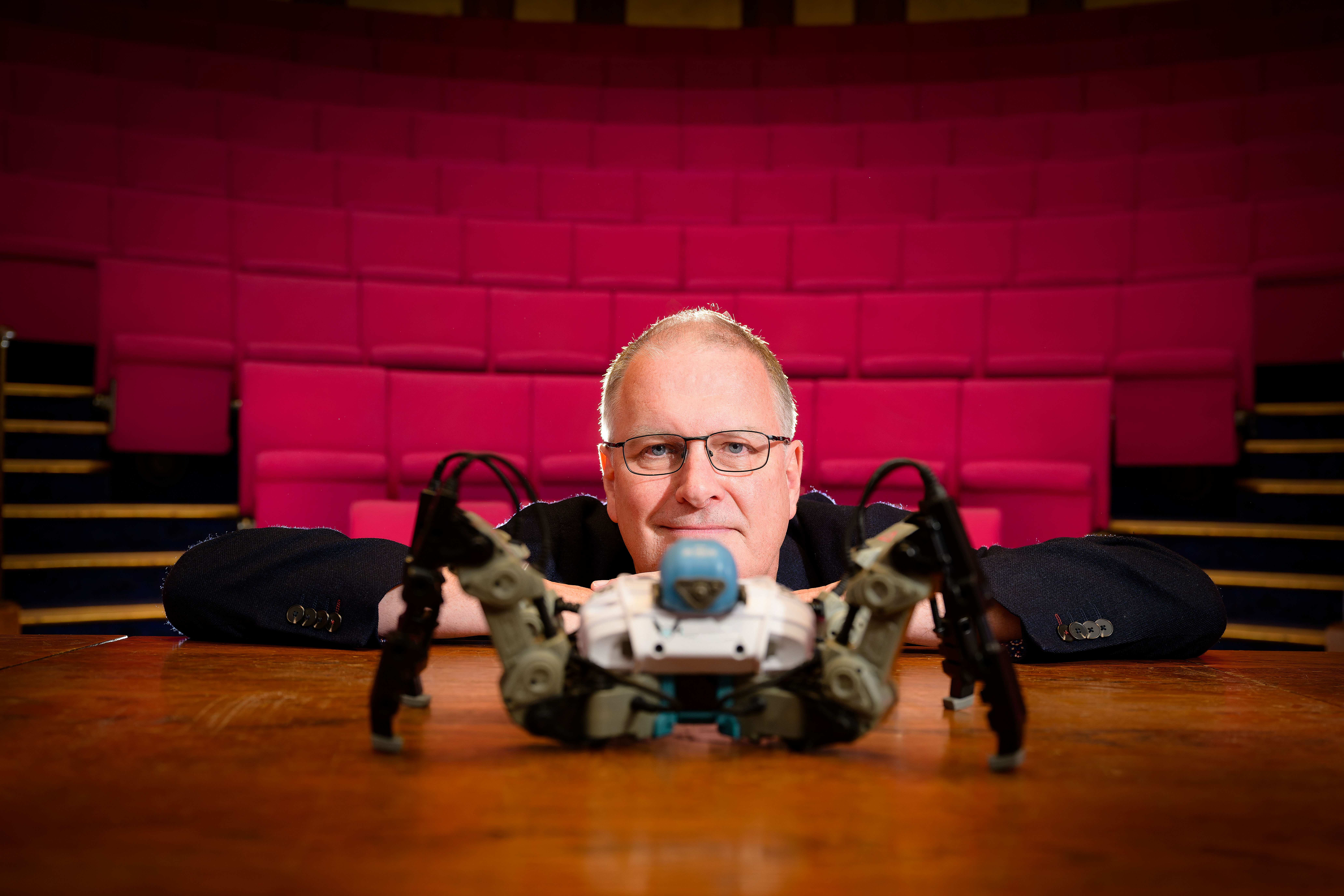 A photograph of Professor Mike Wooldridge leaning on a brown table with a robot in front of him. The background is made up of rows of pink lecture theatre chairs.