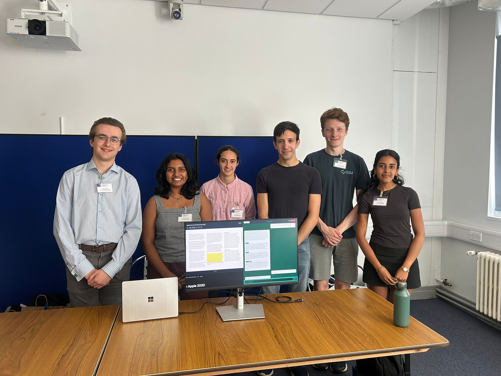 Team 2 smile for the camera. There are six students standing behind a desk with a computer screen showing various documents related to their project.