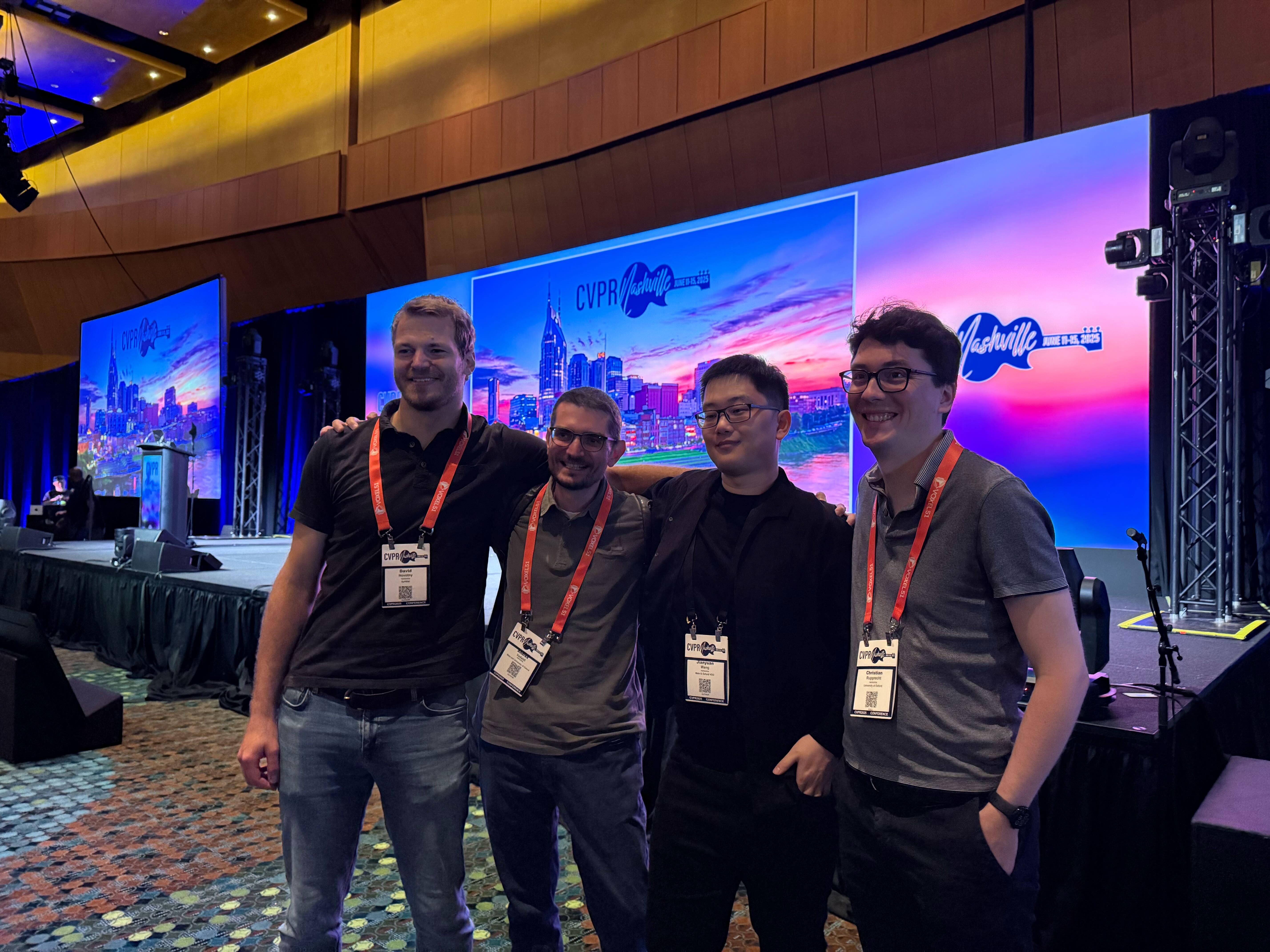 A photograph of four people standing in front of a stage, whilst wearing red lanyards. They are all smiling and looking off to the left of the photo.