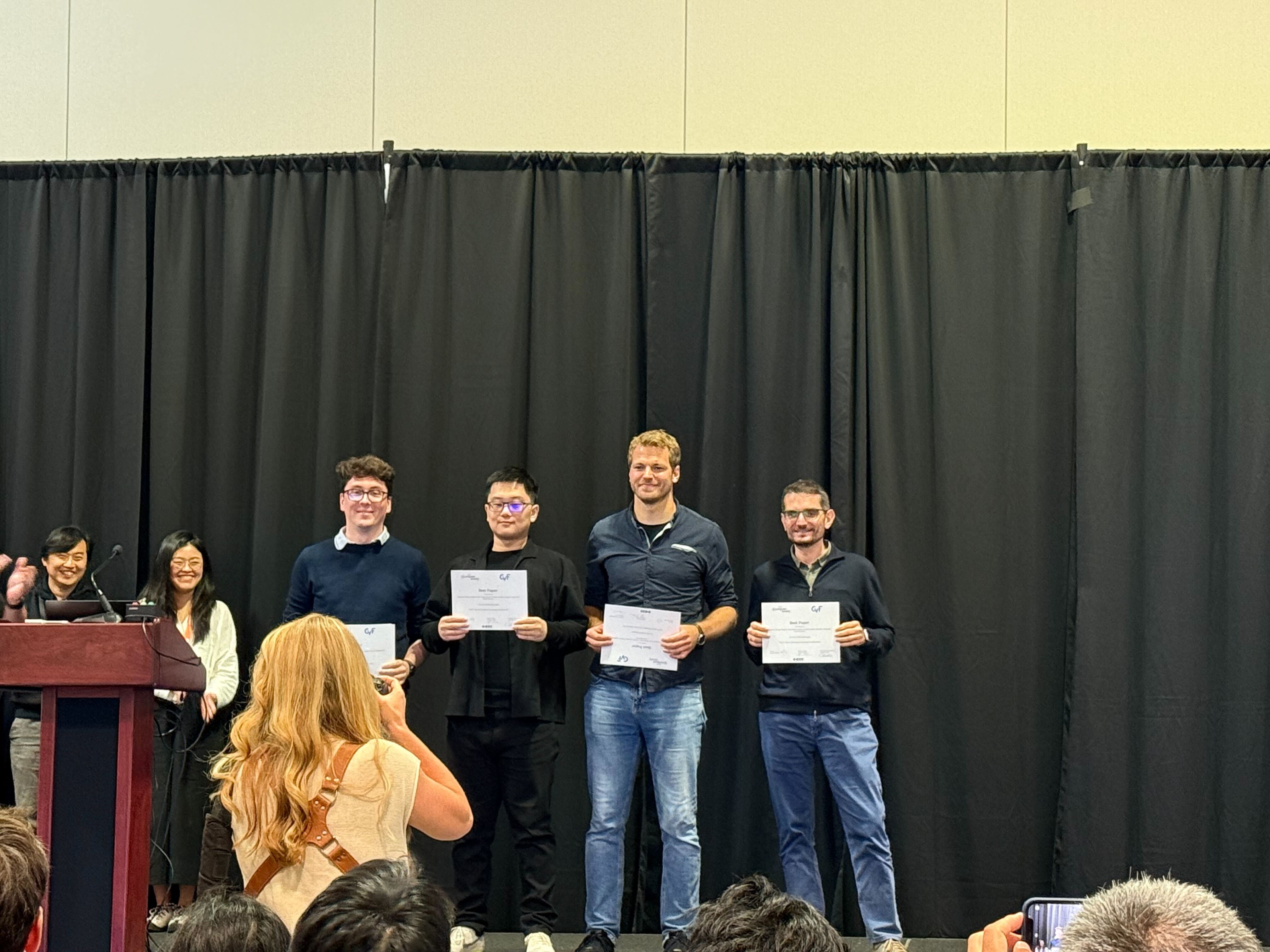 A photograph of four people stood on a stage in front of a black curtain. All four people are holding up white certificates. There are two people at the far left of the stage smiling.