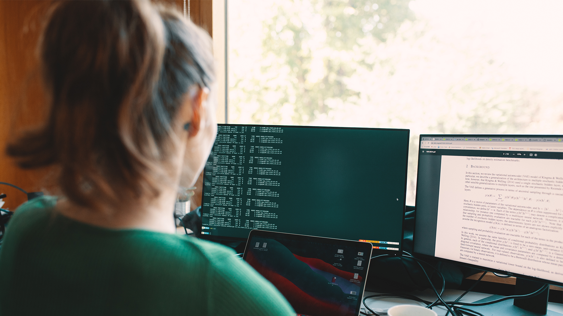 A photograph of the back of a woman's head looking at three computer screens. The computers have text or numbers on them but it is not clearly visible. The woman has straight brown hair tied up and wears a green top. The computer screens are next to a bright window.