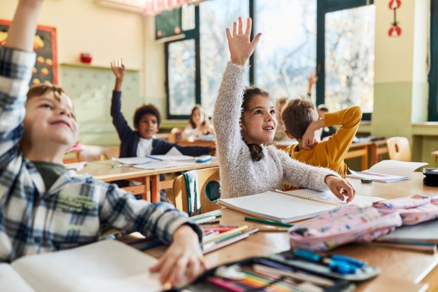 A photograph of a classroom with lots of young children sitting to desks. Two young children in the foreground of the image are smiling and looking forward with their hands up.