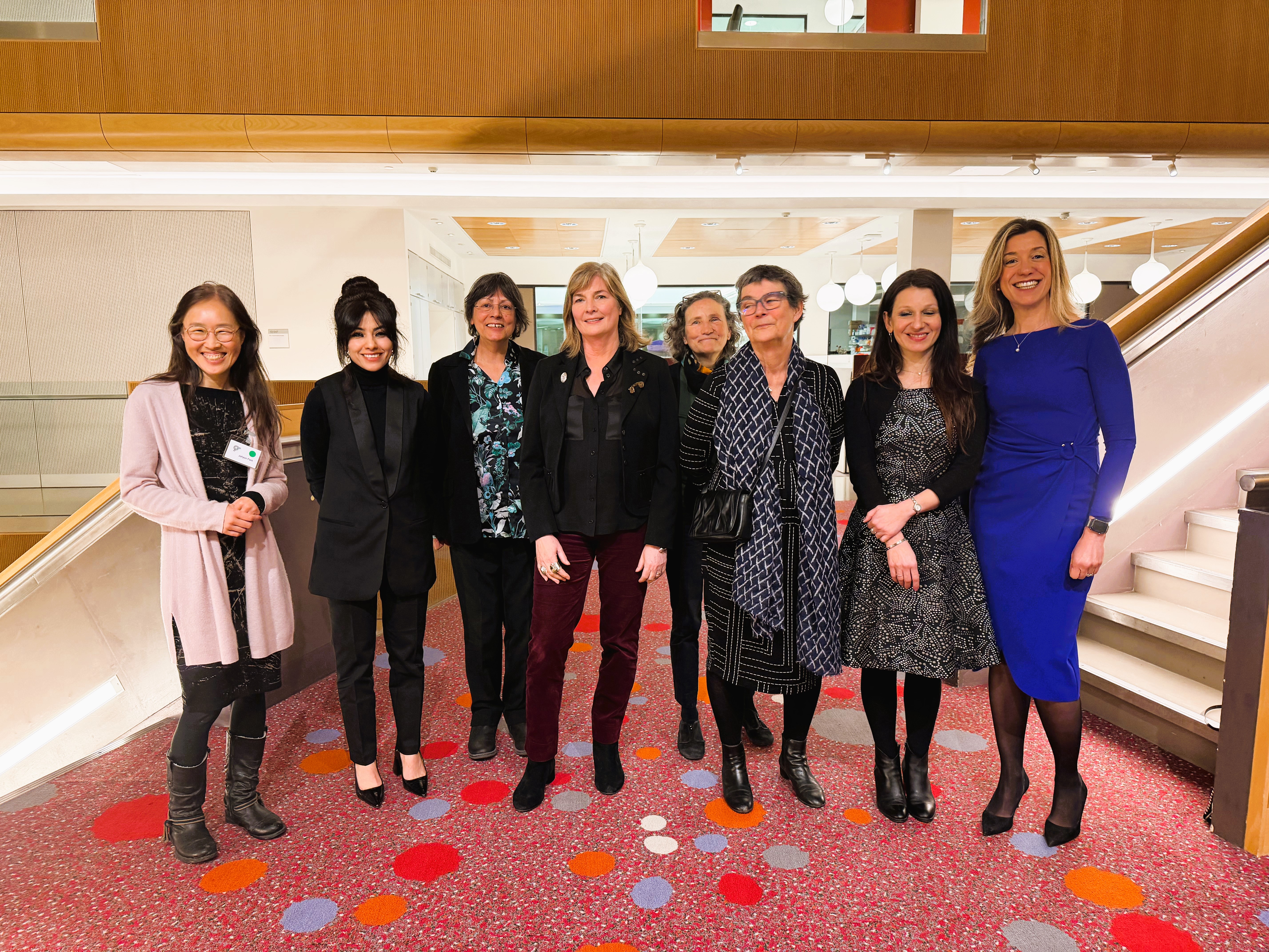 A photo of a group of 8 women standing in smart dress on a red carpet in a stairway of a modern looking building. They are almost all looking at the camera smiling.