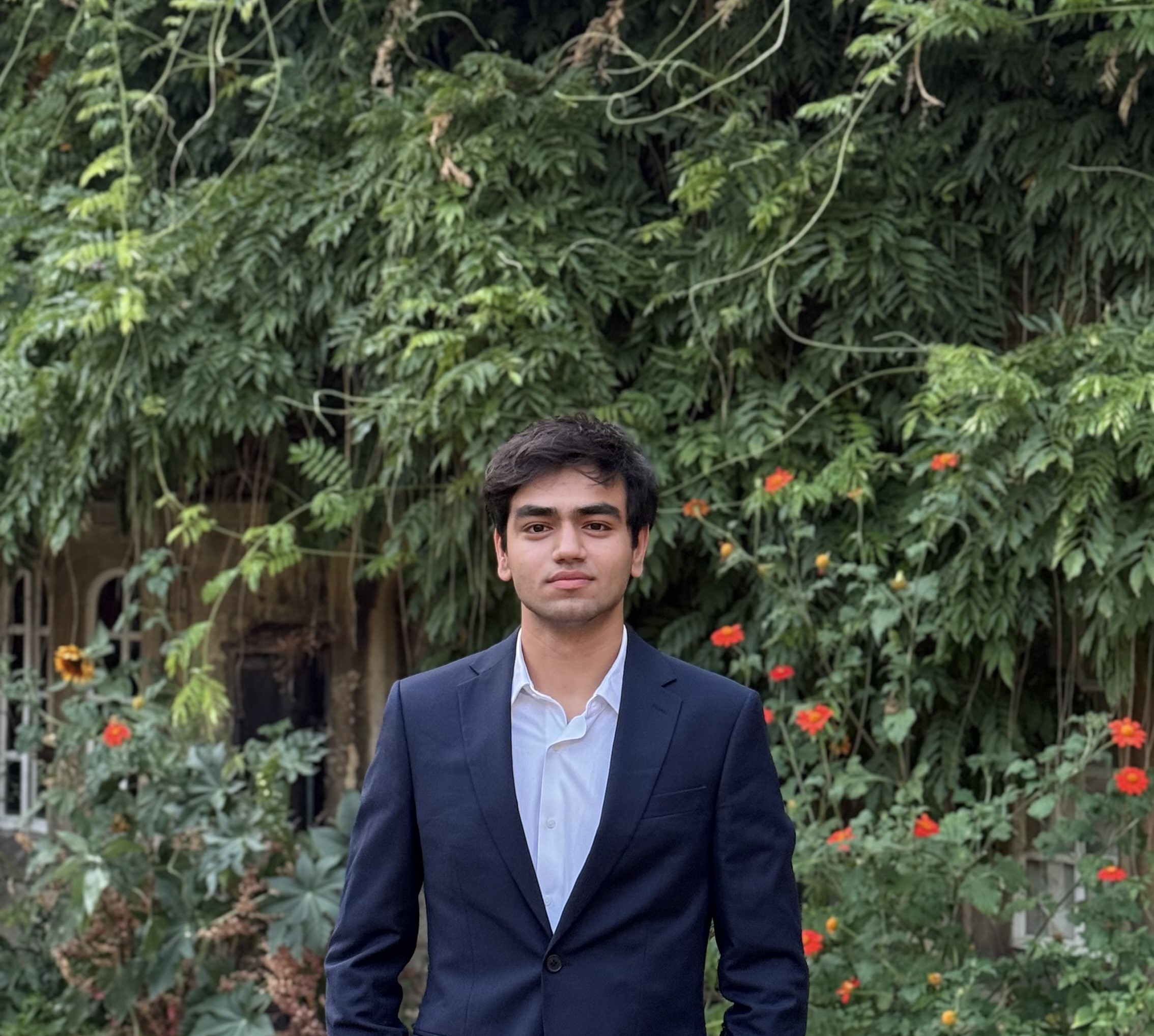 A photo of Tawfeeq Hamayun wearing a smart suit, stood in front of a wall of green plants.